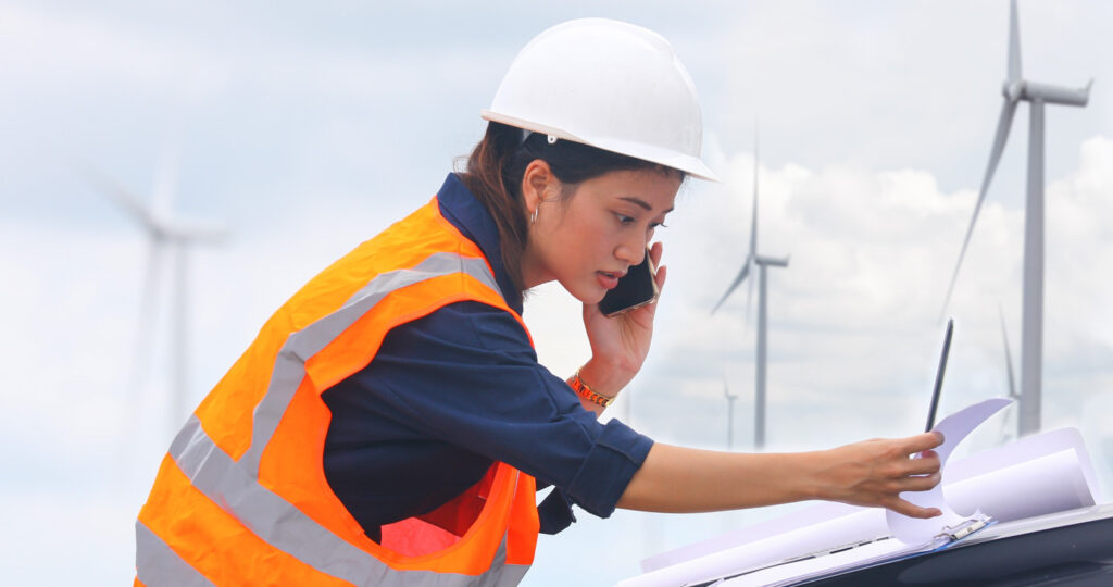 a young woman on a mobile phone and a laptop working on site at a wind turbine farm