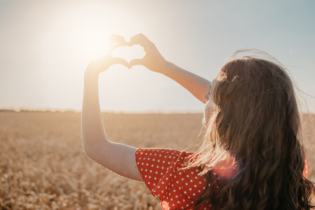 a young woman standing in a corn field making a heart shape with her hands around the sun